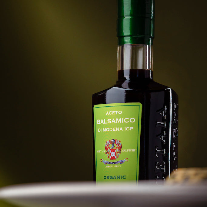 A bottle of Modena IGP balsamic vinegar with a green label and dark glass body, shown on a dark surface with a blurred plate in the foreground.
