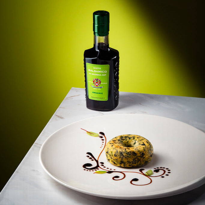 A bottle of organic balsalmic vinegar with a green label and dark glass stands on a white marble surface behind a white plate holding a stuffed bagel with herbs and sauce, set against a yellow-green gradient background.