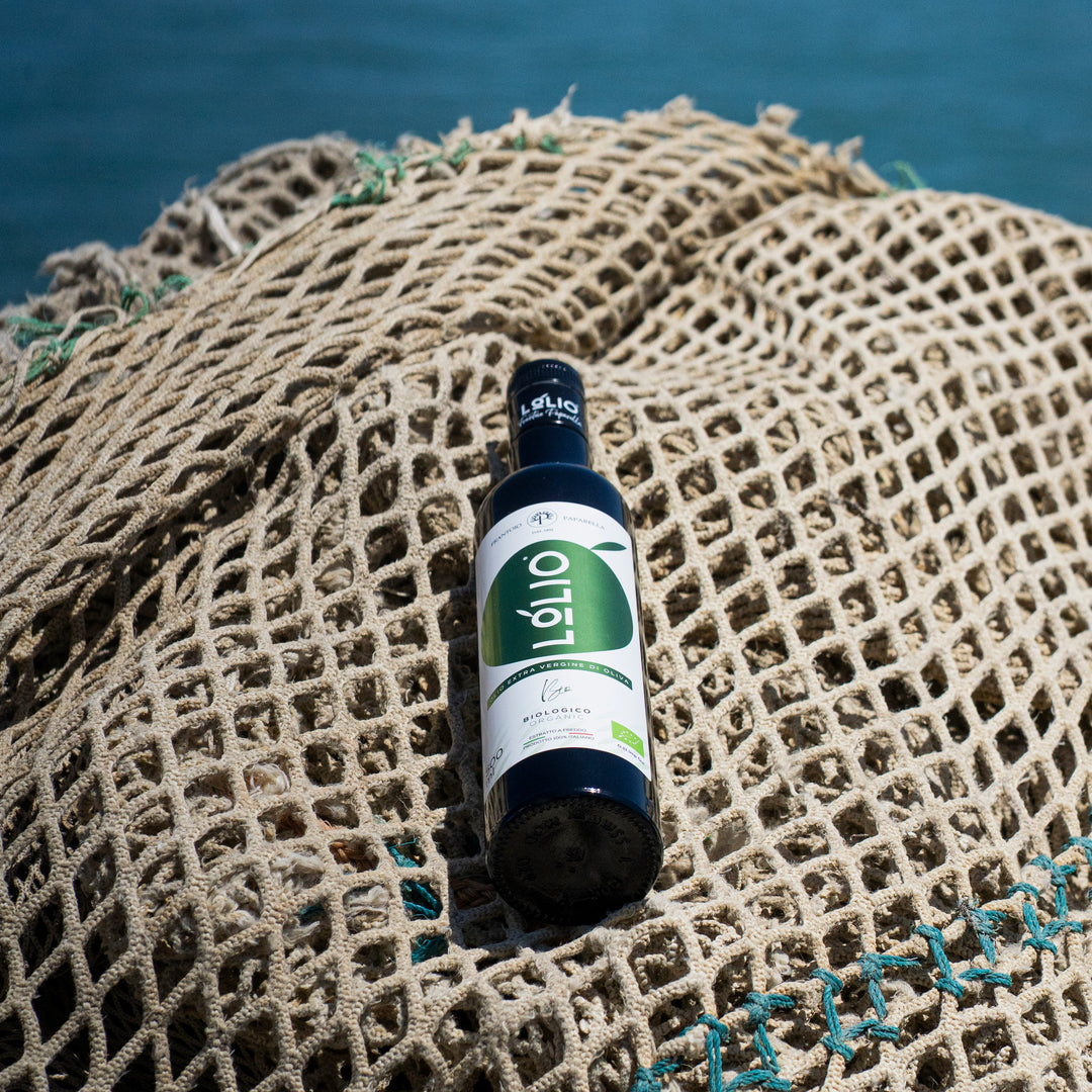 A bottle of olive oil resting on a beige fishing net with blue-green rope, set against a blurred background of deep blue water.