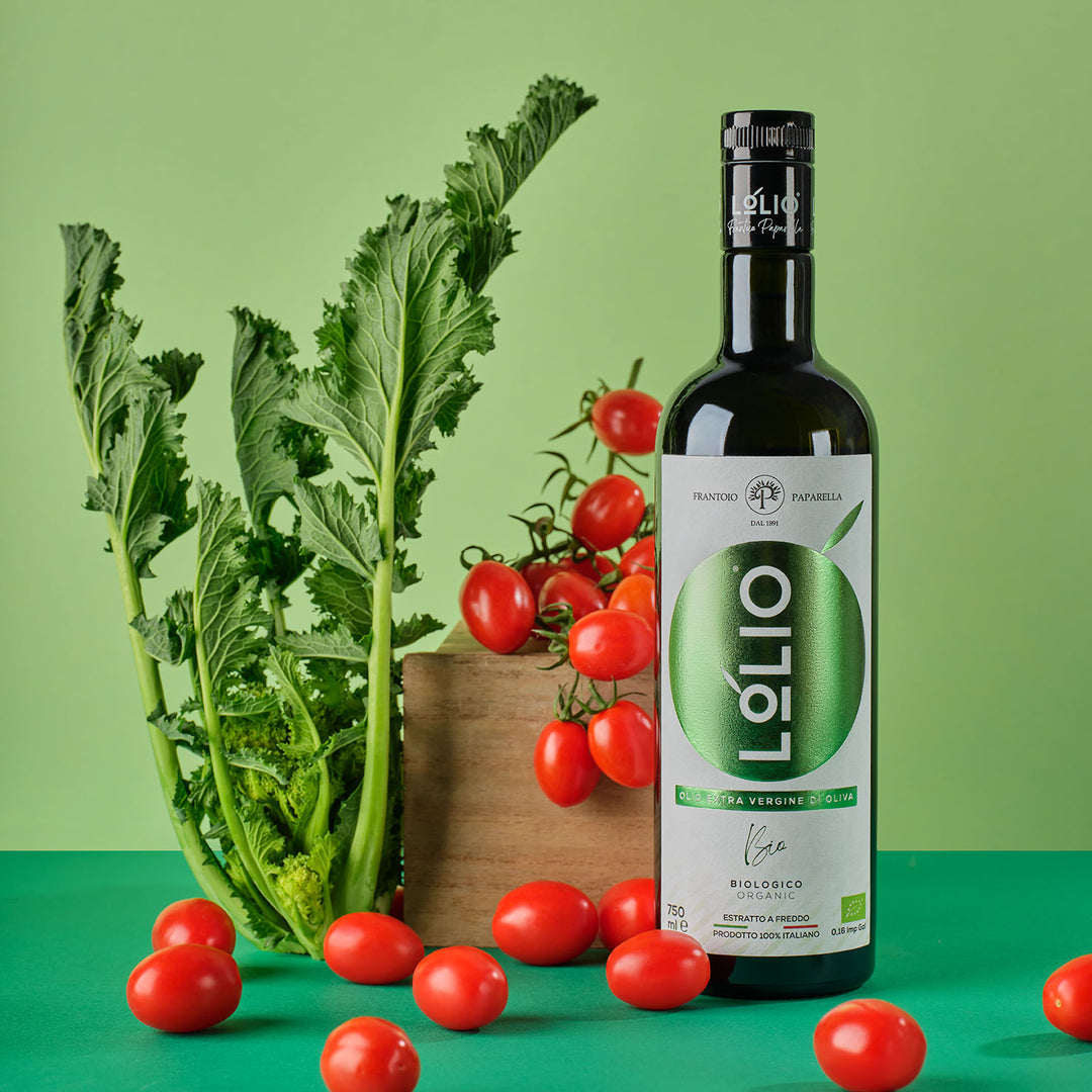 A bottle of Lolio extra virgin olive oil stands on a green surface beside fresh green leafy vegetables, cherry tomatoes on the vine, and scattered cherry tomatoes, against a light green studio background.