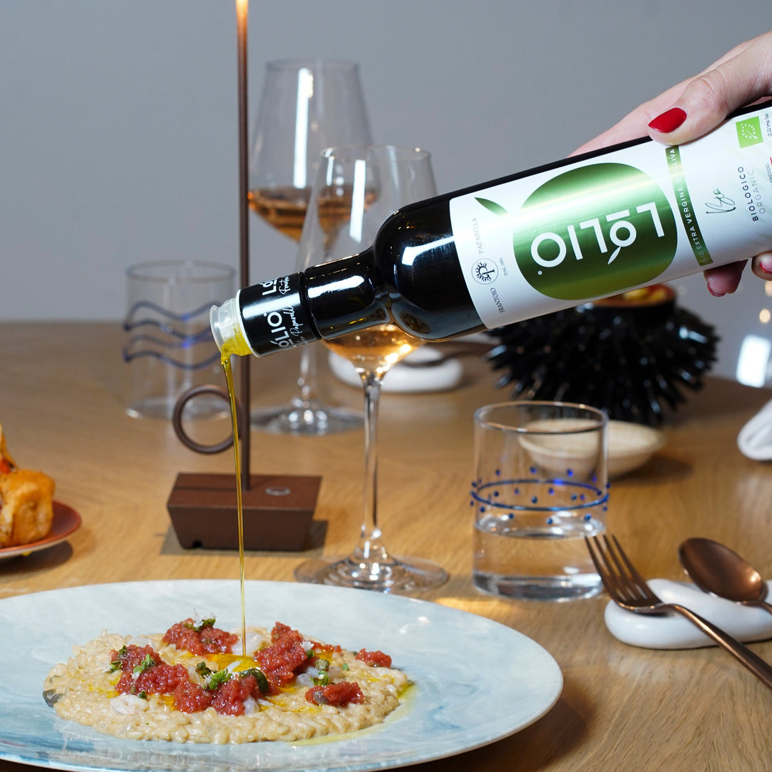 A hand with red nail polish holds a bottle of olive oil and pours golden oil over a plate of risotto with red toppings on a wooden dining table.