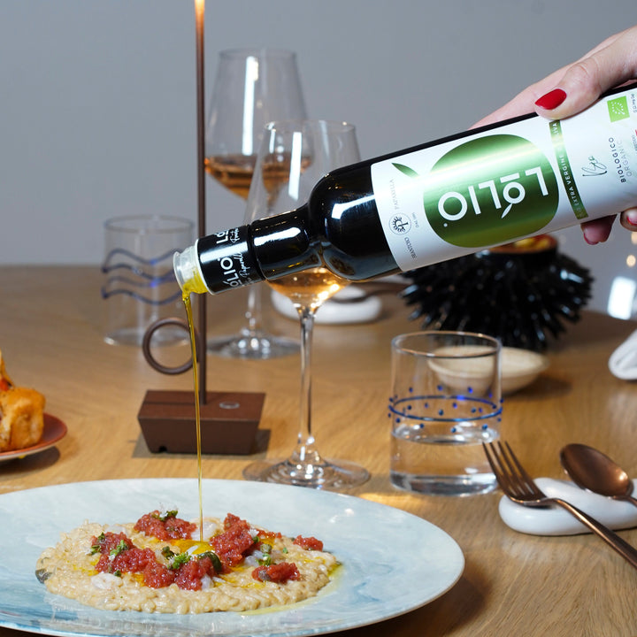 A hand with red nail polish holds a bottle of olive oil and pours golden oil over a plate of risotto with red toppings on a wooden dining table.