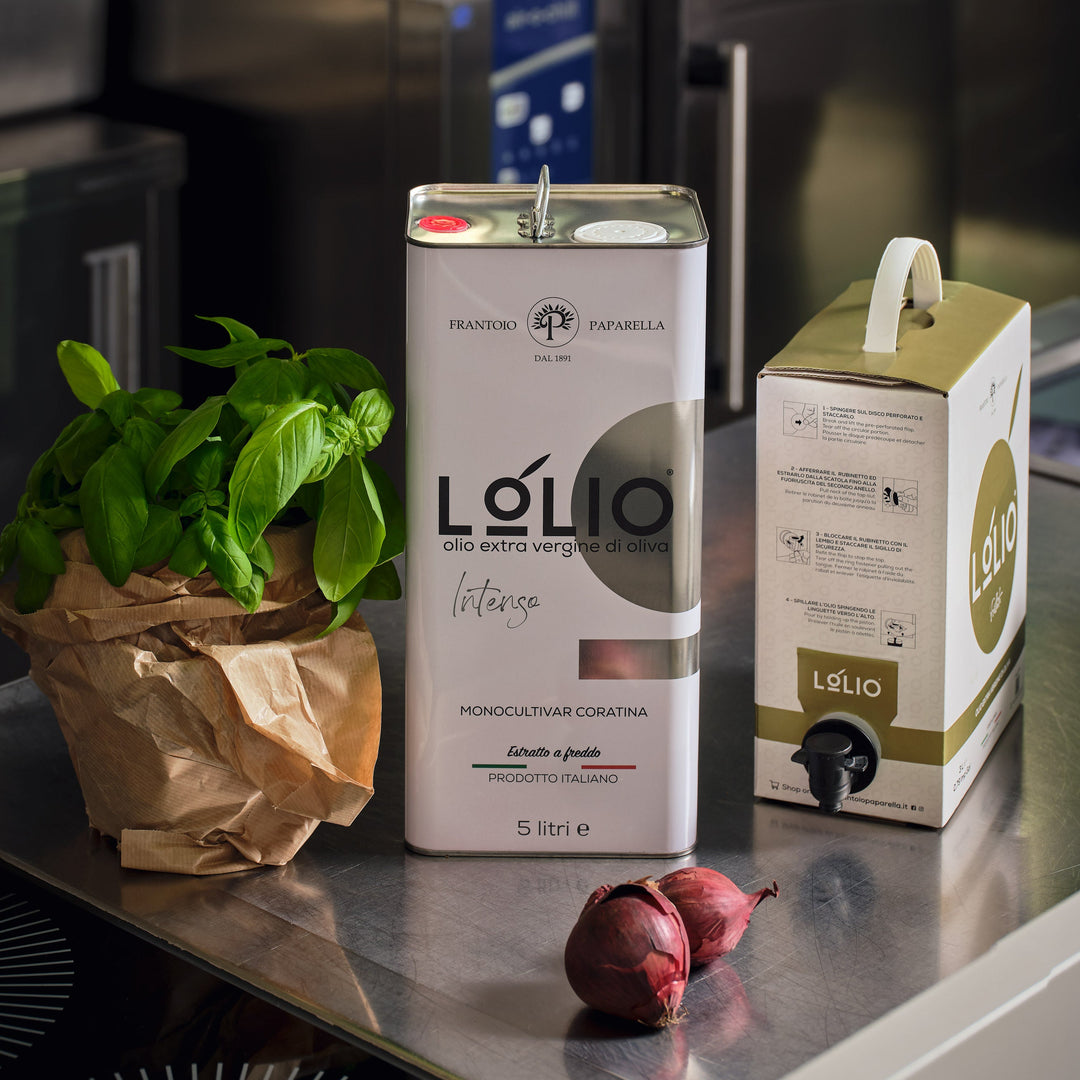 A stainless steel canister of Lólio olive oil stands on a kitchen counter next to a paper bag of fresh basil leaves and two red onions, with a matching Lólio box beside it.
