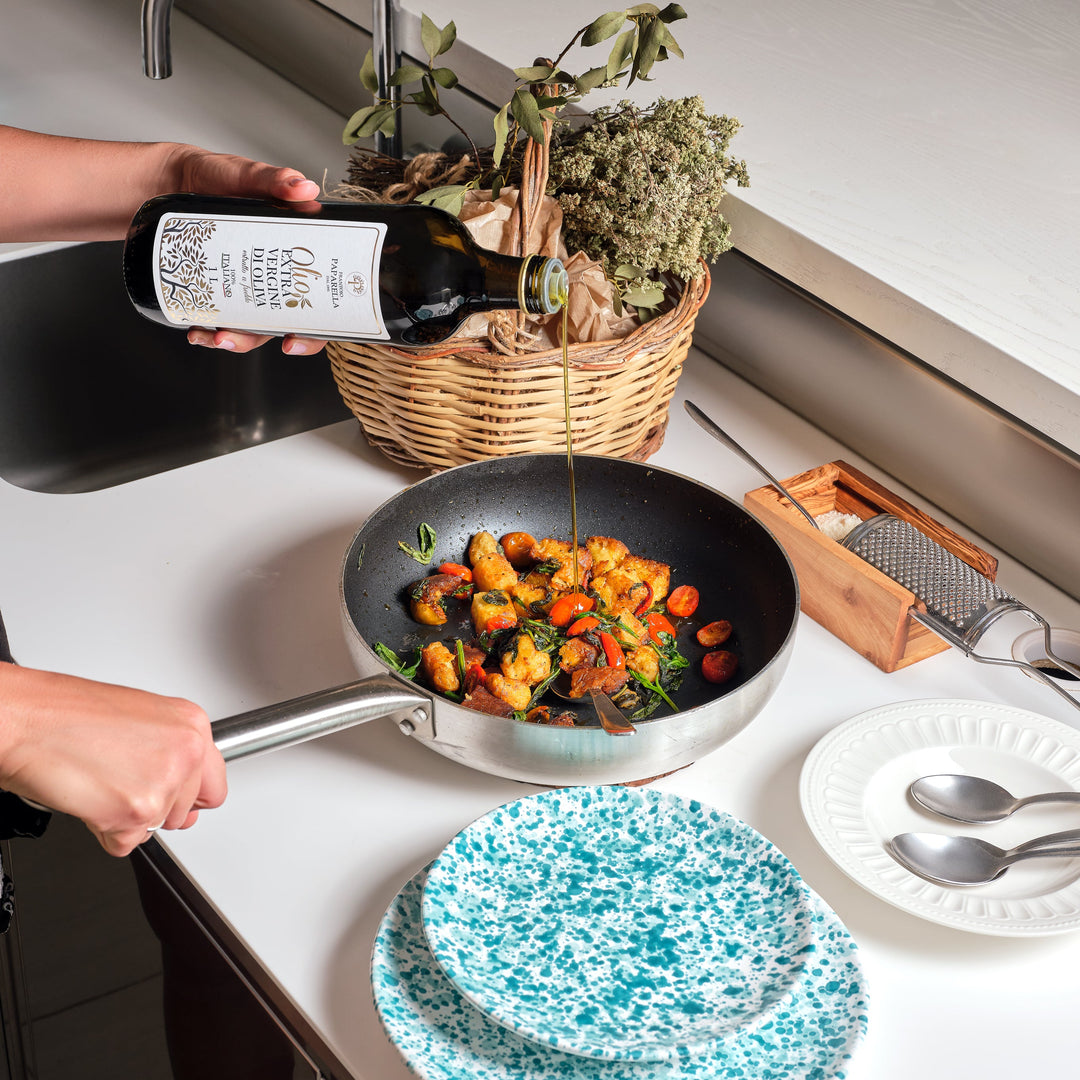 A person holds a bottle of olive oil and pours it into a frying pan on a white kitchen countertop. The scene includes a basket of herbs, a grater, plates, and a sink in a bright kitchen setting.