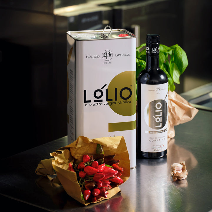 A large white and gold Lolio olive oil canister stands on a dark countertop beside a black bottle of Lolio extra virgin olive oil, with red olives in a gold paper bag, garlic cloves, and green basil leaves in a kitchen setting.