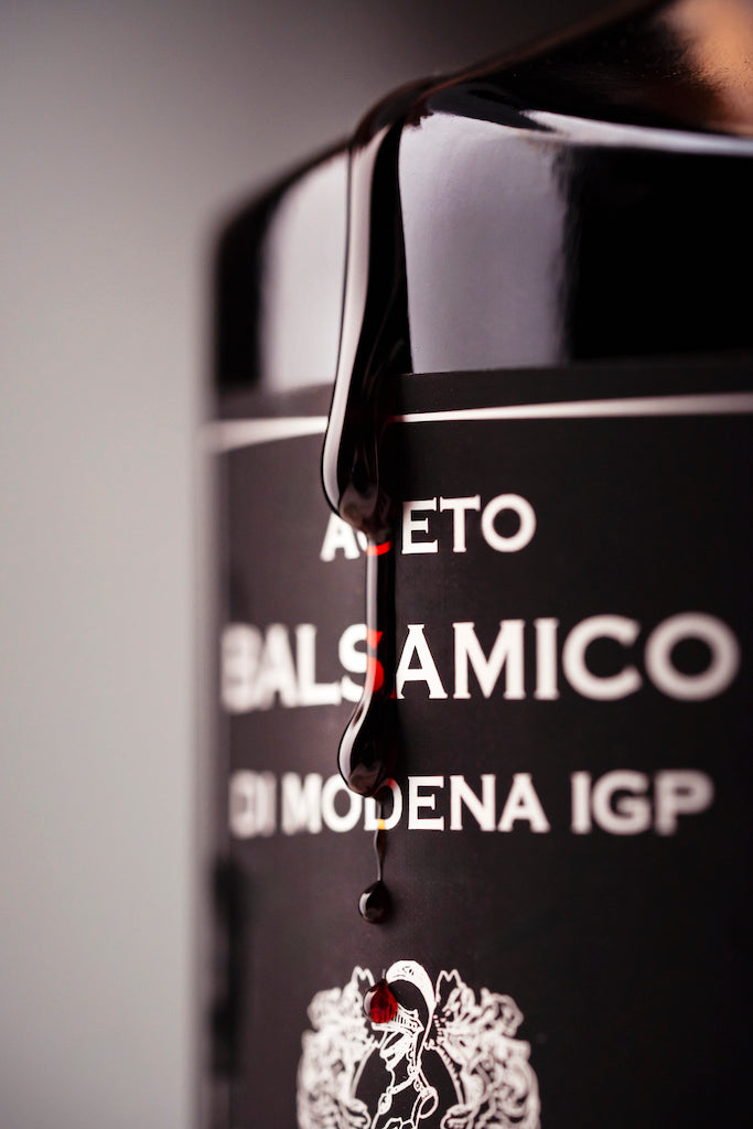 Close-up of a black bottle labeled 'ACETO BALSAMICO DI MODENA IGP' with glossy dark syrup dripping from the top against a soft gray background.