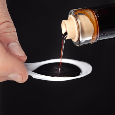 A close-up of a hand holding a white spoon while a bottle pours a dark liquid, platinum balsamic vinegar, onto the spoon against a black background.