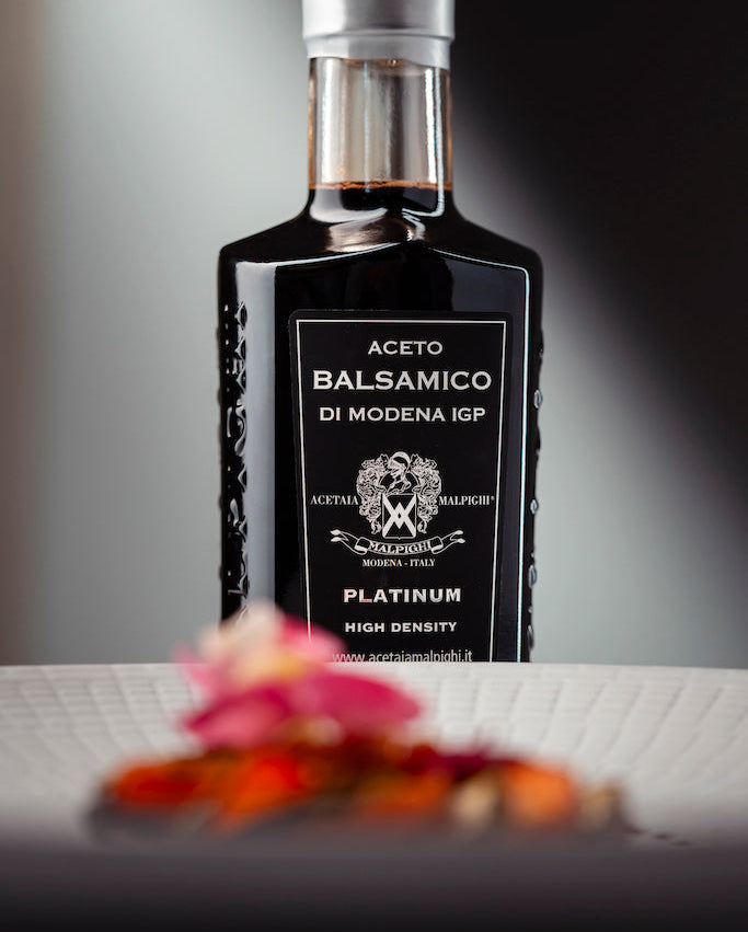 A bottle of Aceto Balsamico di Modena IGP Platinum high density balsamic vinegar, featuring a black label and silver cap, set against a dark background with blurred pink flower petals in the foreground.