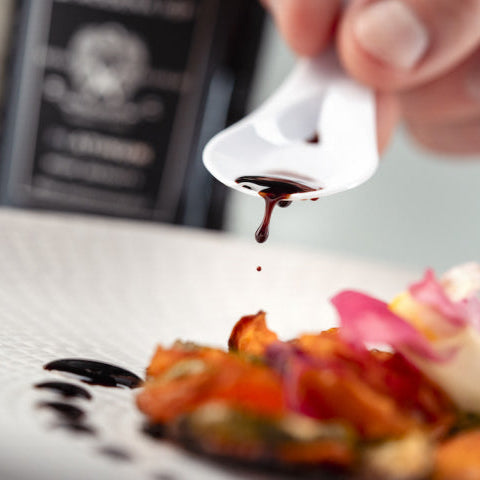 A close-up of a hand holding a white spoon and drizzling dark balsamic sauce over a plated gourmet salad or appetizer, with a blurred bottle in the background.