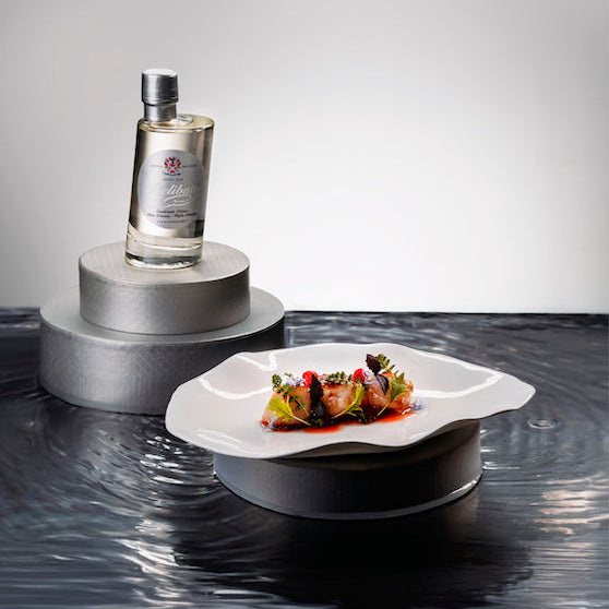 A clear glass bottle with a silver cap displayed on a metallic pedestal, alongside a white plate of food, set on a dark reflective surface against a light gray background.
