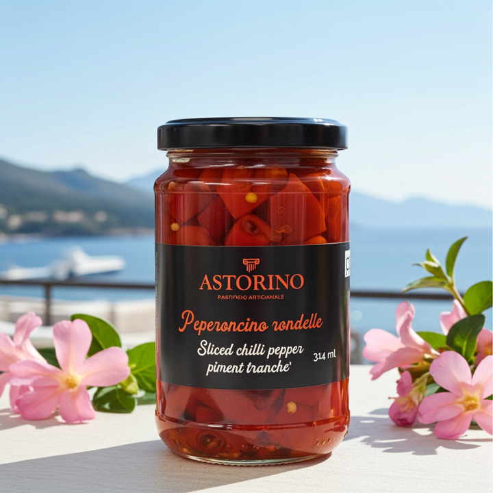 A jar of Astorino sliced chili pepper (peperoncino rondello) sits on a table outdoors with a blurred coastal background and pink flowers framing the jar.
