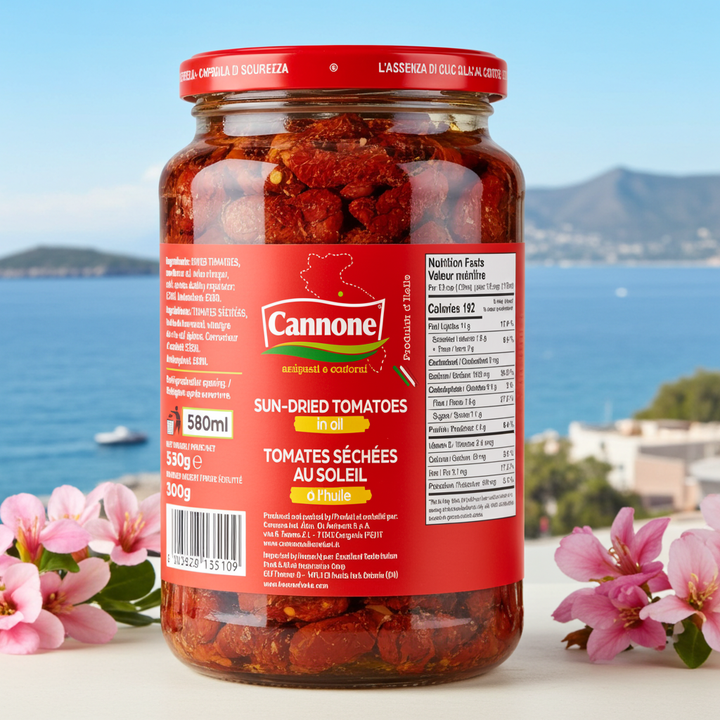 A 580ml glass jar of Cannone Sun-Dried Tomatoes in oil, set against a scenic Mediterranean background with blue water and mountains, framed by pink flowers at the base.