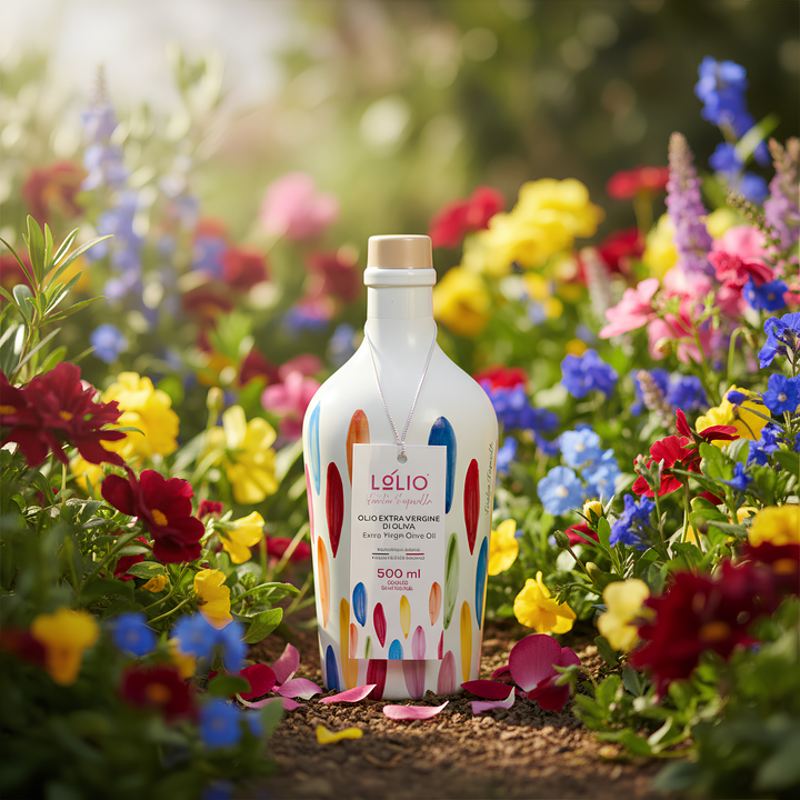 A white bottle of Lolio olive extra virgin oil stands in a garden surrounded by colorful flowers. The bottle features a white label with a colorful abstract pattern of red, blue, yellow, and orange shapes.