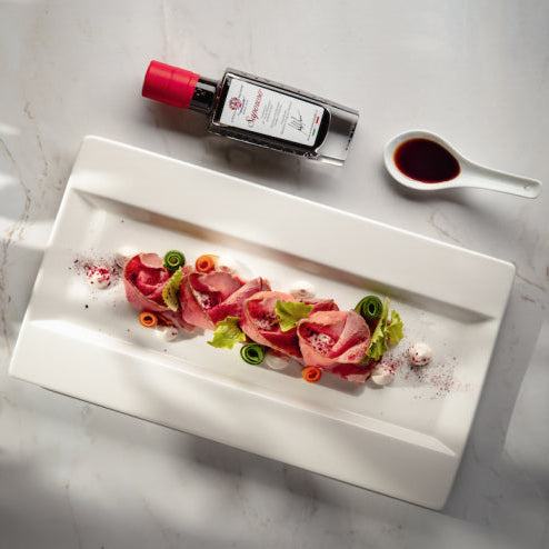 A styled flat-lay of a white rectangular platter holding thinly sliced pink cured meat with green and orange garnish, set on a light marble surface. A bottle with a red cap, a small white spoon with dark sauce, and a napkin with green leaves are arranged around it.