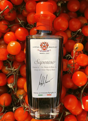 A bottle of Saparoso Dressing with Balsamic Vinegar of Modena  centered against a dense background of fresh red cherry tomatoes. The bottle has a clear glass body showing the dark liquid inside, a white label with the brand name, and a bright orange-red cap.