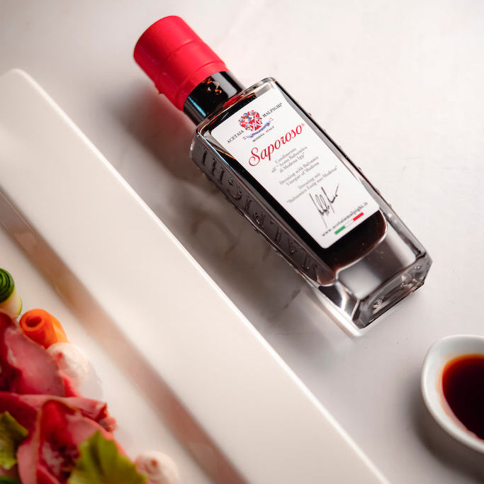A bottle of Saparoso Dressing with Balsamic Vinegar of Modena  with a red cap and white label, styled on a white marble surface next to a plate of food.