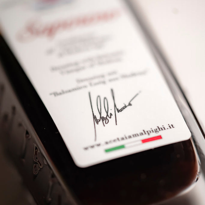 A close-up, angled shot of a dark glass bottle containing a dark red liquid, featuring a white label with a signature and the website www.acetiamalpigh.