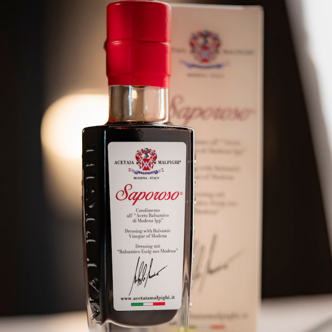 A bottle of Saporoso vinegar with a red cap and white label, standing in front of its branded box on a blurred tabletop setting.