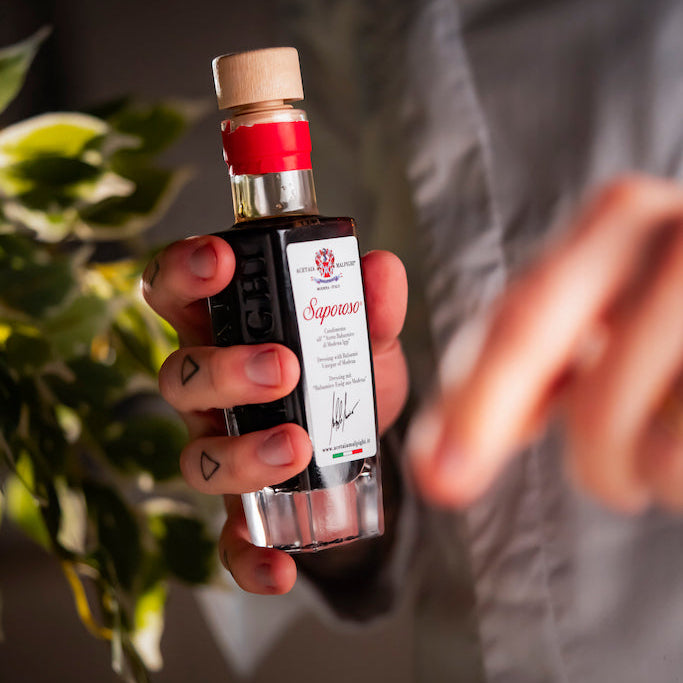 A hand holding a small bottle of Saparoso Dressing with Balsamic Vinegar of Modena, with a person in a gray shirt blurred in the background and green leaves on the left.
