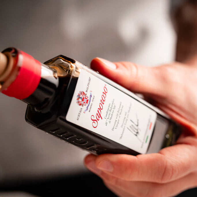 A close-up of a person's hands holding a bottle of Saparoso Dressing with Balsamic Vinegar of Modena over a plate of food, with the brand name clearly visible on the label.