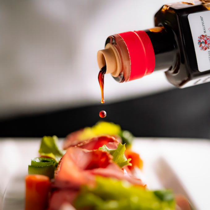 A close-up of a hand holding a dark bottle with a red cap, pouring a glossy dark sauce over a plated salad with sliced red meat, green lettuce, and orange carrot pieces.