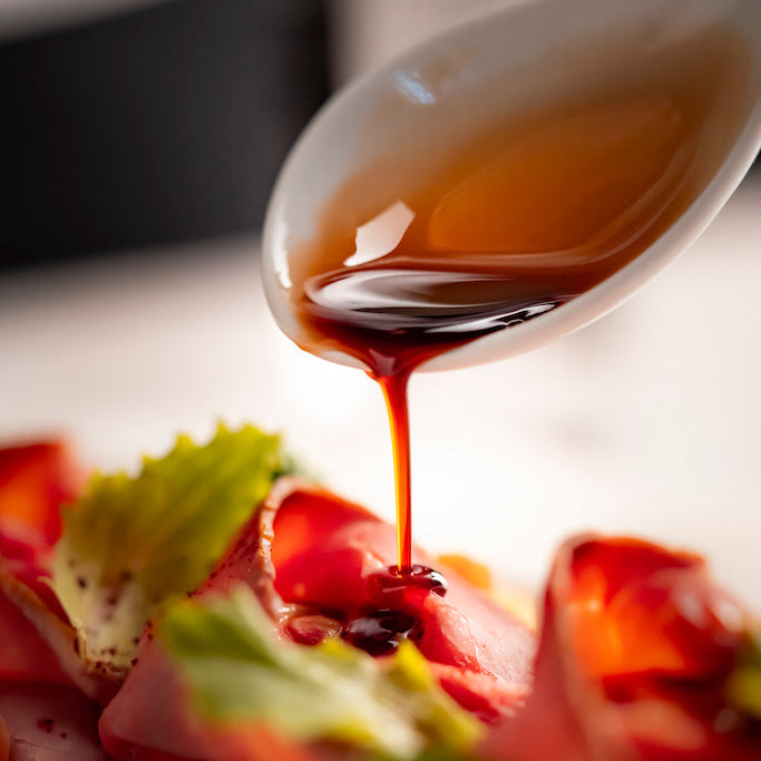 A close-up shot of a glossy amber-brown sauce being drizzled from a white spoon over a plated food dish with sliced red meat, green lettuce, and a small green vegetable on a white plate.
