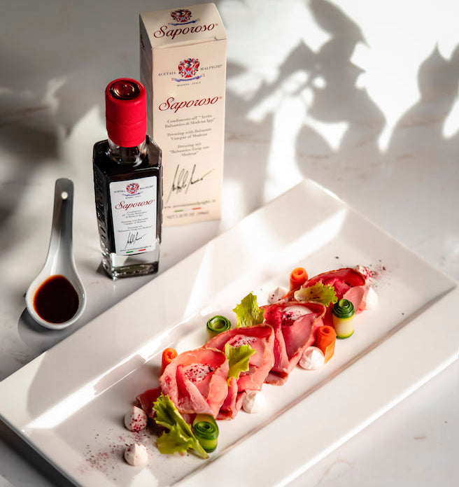 A styled food presentation featuring a long white rectangular platter with a gourmet dish of thinly sliced pink meat, green herbs, and orange carrot pieces, alongside a bottle and box of Saponekó wine in a clear glass, and a small spoon of dark red sauce on a white marble surface with leaf shadows.