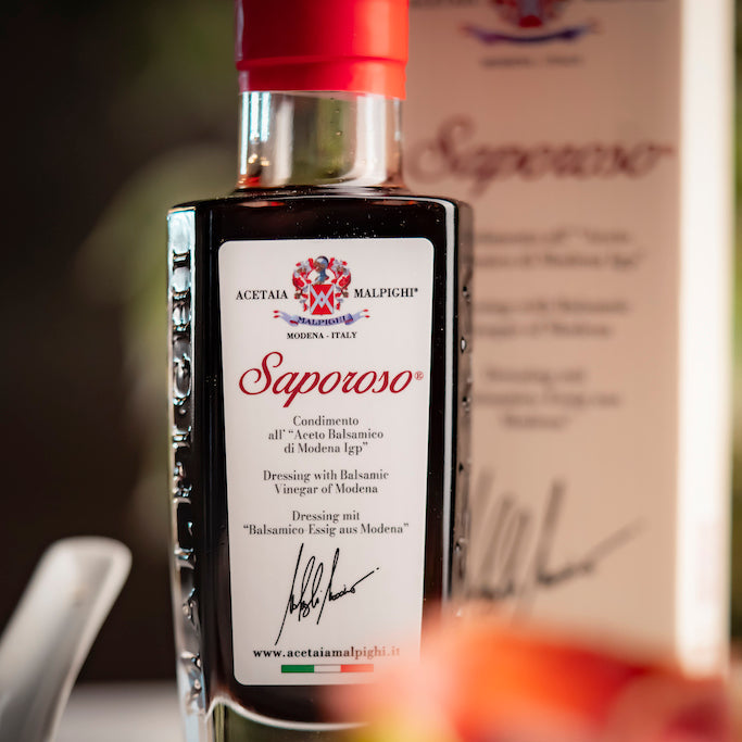 A bottle of Saparoso Dressing with Balsamic Vinegar of Modena  featuring a dark glass body, a white label with red and black text, and a red cap, set against a blurred background with a matching box.