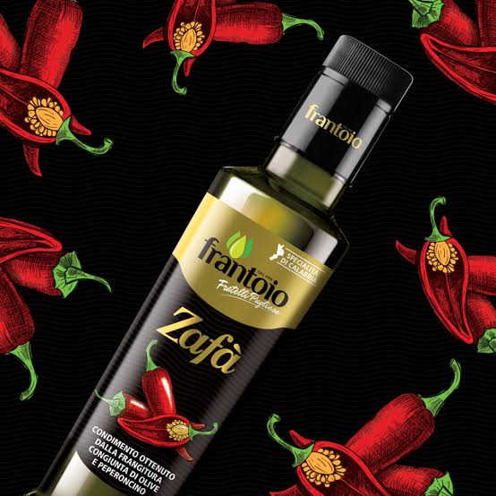 A bottle of Frantoio Zafa extra virgin olive oil, styled diagonally on a black background decorated with red chili pepper illustrations. The bottle features a black cap and a label with a gold top section and red chili pepper graphics.