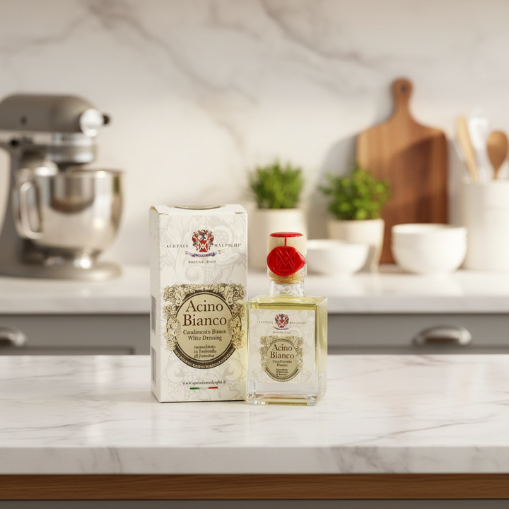 A bottle of Acino Bianco White Dressing and its matching box, both featuring a vintage-style cream label with a red crest, displayed on a marble kitchen countertop with a blurred kitchen background.