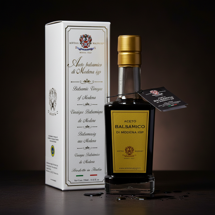 A bottle of Aceto Balsamico di Modena 109P balsamic vinegar with a gold cap and yellow label, standing next to its white branded box on a dark surface against a dark brown background.
