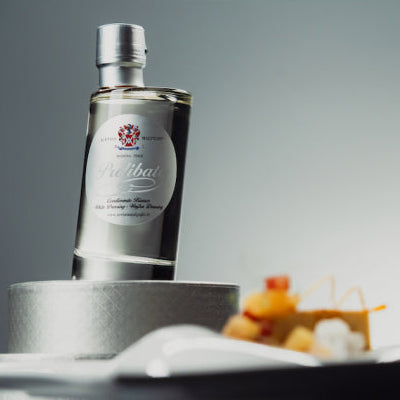 A clear glass bottle of acetaia malpighiWhite Dressing with a silver cap, displayed on a metallic stand with a blurred background and small food props in the foreground.