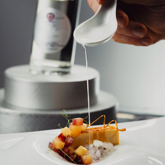 A close-up of a hand holding a white ceramic spoon and pouring a thin white sauce over a plated gourmet dish. In the softly blurred background, a glass bottle with a silver cap sits on a metallic stand against a dark gray backdrop.