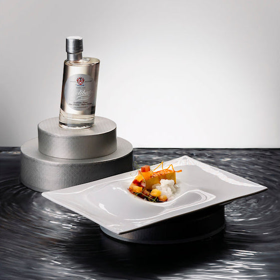 A clear glass bottle with a silver cap and label stands on a metallic pedestal, next to a white square plate holding a small gourmet food garnish, set against a dark reflective surface.