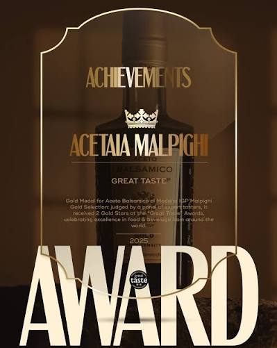 A dark brown glass award plaque with gold lettering and a gold crown emblem, featuring the text 'Achievements Acetaia Malpighi Balsamico Great Taste 2025' and a large 'AWARD' at the bottom.