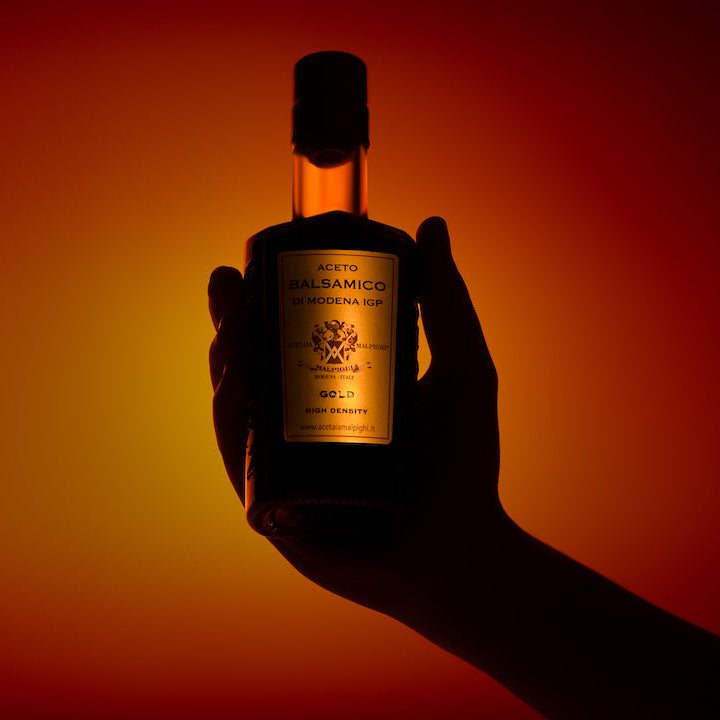 A hand holding a bottle of Aceto Malpighi Gold balsamic vinegar against a dramatic red and orange gradient background.