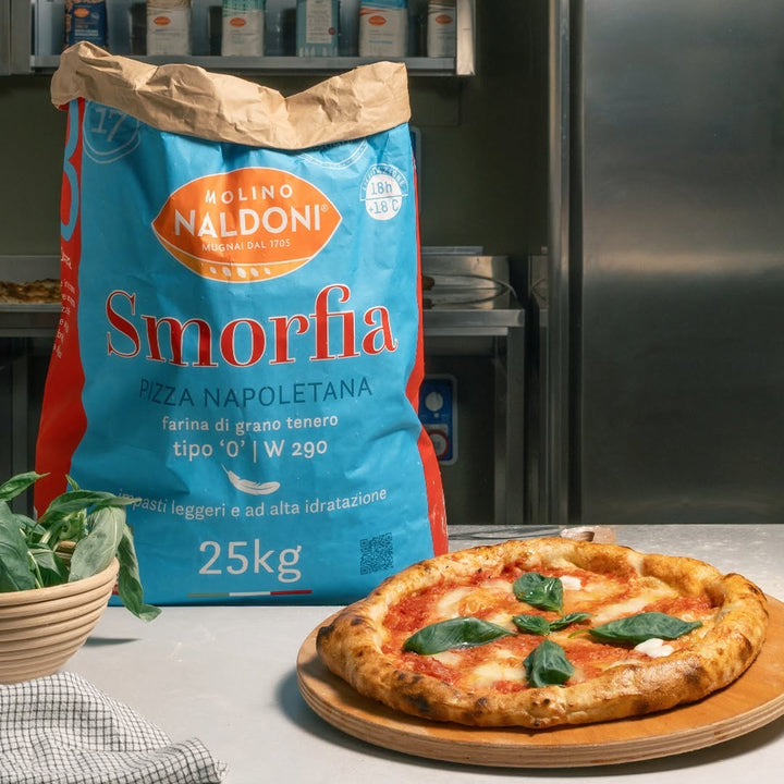 A 25kg bag of Molino Naldoni Smorfia pizza flour stands on a kitchen counter next to a freshly baked pizza with basil leaves on a wooden board, with a stainless steel refrigerator and shelves in the background.