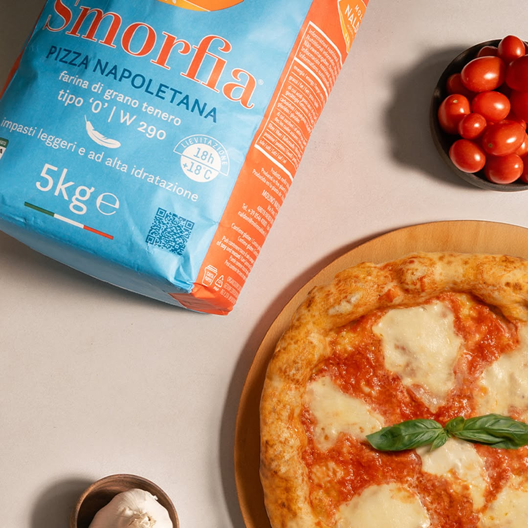 A 5kg bag of Smorfia pizza flour styled on a light beige surface next to a freshly baked pizza on a wooden board, with small bowls of cherry tomatoes and garlic.