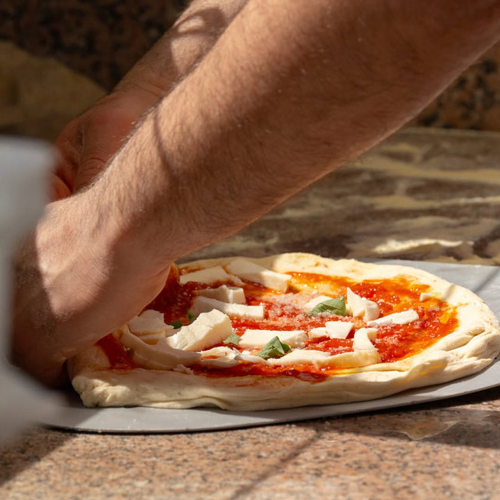 A close-up of a hand placing an uncooked pizza onto a metal peel on a stone pizza oven floor. The pizza is topped with tomato sauce, white mozzarella cheese, and small green basil leaves.